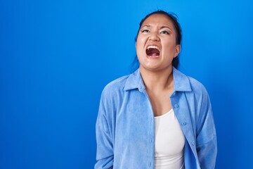 Canvas Print - Asian young woman standing over blue background angry and mad screaming frustrated and furious, shouting with anger. rage and aggressive concept.