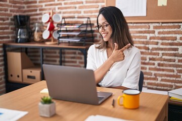 Poster - Hispanic young woman working at the office wearing glasses smiling cheerful pointing with hand and finger up to the side