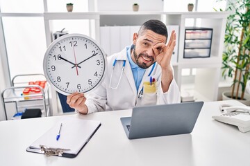 Wall Mural - Hispanic doctor man holding clock at the clinic smiling happy doing ok sign with hand on eye looking through fingers