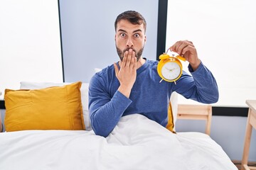 Wall Mural - Handsome hispanic man in the bed holding alarm clock covering mouth with hand, shocked and afraid for mistake. surprised expression