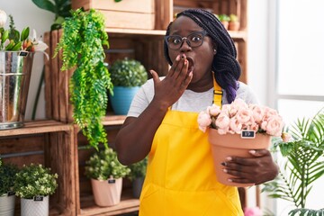 Wall Mural - Young african woman working at florist shop holding plant covering mouth with hand, shocked and afraid for mistake. surprised expression