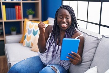 Poster - Young african woman using touchpad sitting on the sofa screaming proud, celebrating victory and success very excited with raised arm