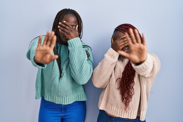 Two african woman standing over blue background covering eyes with hands and doing stop gesture with sad and fear expression. embarrassed and negative concept.