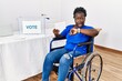 Young african woman sitting on wheelchair voting putting envelop in ballot box looking at the watch time worried, afraid of getting late