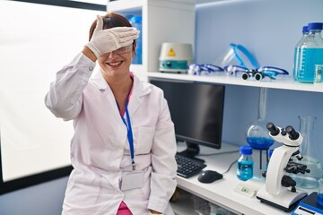 Wall Mural - Young brunette woman working at scientist laboratory smiling and laughing with hand on face covering eyes for surprise. blind concept.