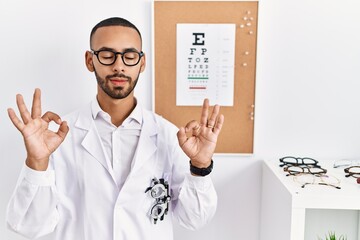 Canvas Print - African american optician man standing by eyesight test relaxed and smiling with eyes closed doing meditation gesture with fingers. yoga concept.