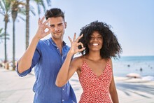 Young Interracial Couple Outdoors On A Sunny Day Smiling Positive Doing Ok Sign With Hand And Fingers. Successful Expression.