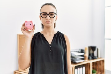 Poster - Young brunette woman holding piggy bank at the office thinking attitude and sober expression looking self confident