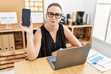 Poster - Young brunette woman holding smartphone at the office thinking attitude and sober expression looking self confident