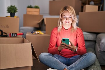 Canvas Print - Young blonde woman using smartphone sitting on floor at new home