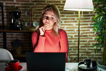 Canvas Print - Blonde woman using laptop at night at home with hand on chin thinking about question, pensive expression. smiling with thoughtful face. doubt concept.