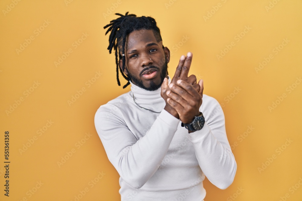 African man with dreadlocks wearing turtleneck sweater over yellow background holding symbolic gun with hand gesture, playing killing shooting weapons, angry face