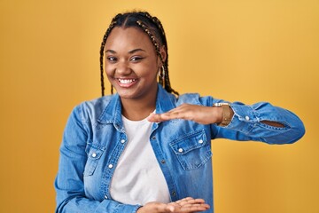 Canvas Print - African american woman with braids standing over yellow background gesturing with hands showing big and large size sign, measure symbol. smiling looking at the camera. measuring concept.