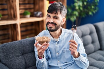 Sticker - Young hispanic man with beard eating healthy whole grain cereals winking looking at the camera with sexy expression, cheerful and happy face.