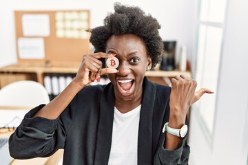 Poster - African young woman holding bitcoin on eye at the office pointing thumb up to the side smiling happy with open mouth