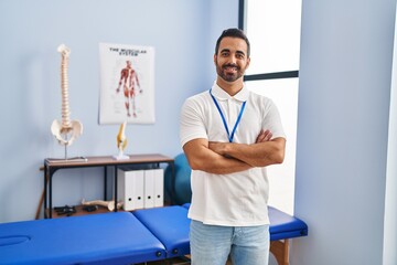 Wall Mural - Young hispanic man physiotherapist standing with arms crossed gesture at rehab clinic