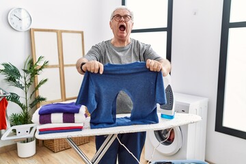 Poster - Senior caucasian man ironing holding burned iron shirt at laundry room angry and mad screaming frustrated and furious, shouting with anger looking up.