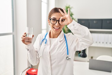 Poster - Young brunette doctor woman holding glass of water smiling happy doing ok sign with hand on eye looking through fingers