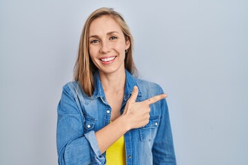 Sticker - Young blonde woman standing over blue background cheerful with a smile on face pointing with hand and finger up to the side with happy and natural expression