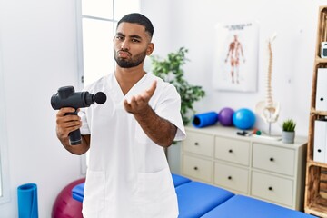 Sticker - Young indian physiotherapist holding therapy massage gun at wellness center looking at the camera blowing a kiss with hand on air being lovely and sexy. love expression.