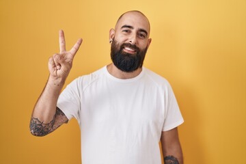 Canvas Print - Young hispanic man with beard and tattoos standing over yellow background showing and pointing up with fingers number two while smiling confident and happy.