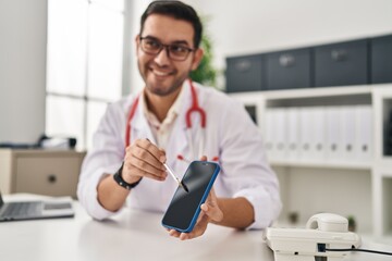 Poster - Young hispanic man wearing doctor uniform showing medical smartphone app at clinic