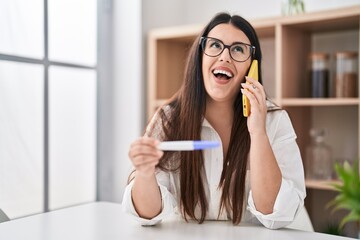 Poster - Young brunette woman holding pregnancy test result speaking on the phone smiling and laughing hard out loud because funny crazy joke.