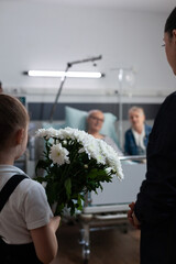 Wall Mural - Elderly couple being visited by daughter, granddaughter in geriatric clinic rest room. Little girl carrying flowers gift to recovering grandfather lying in hospital bed.