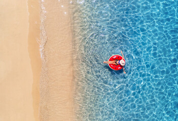 Poster - Aerial view of a woman in hat swimming with red swim ring in blue sea at sunrise in summer. Tropical landscape with girl, clear water, waves, sandy beach. Top view. Vacation. Sardinia island, Italy	