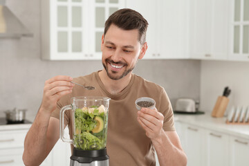 Canvas Print - Happy man adding chia seeds into blender with ingredients for smoothie in kitchen