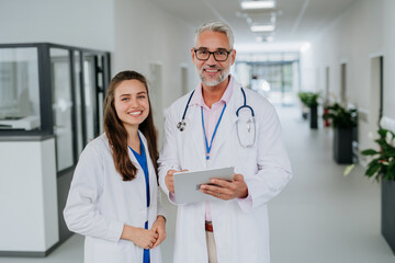 Portrait of elderly doctor with his younger colleague at hospital corridor. Health care concept.