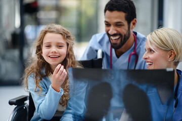 Wall Mural - Young multiracial doctor with his colleague showing x-ray image of lungs to little girl on wheelchair.