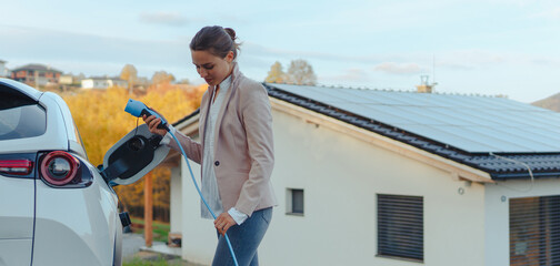 Wall Mural - Young woman charging her electric car in home, sustainable and economic transportation concept.