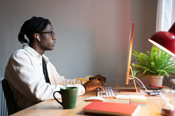 Wall Mural - Focused concentrated African man freelance web developer wearing spectacles writing code using computer, sitting at cozy home office. Smart skilled black guy, computer programmer coding remotely