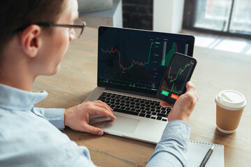 Rare view cropped closeup picture of intelligent successful crypto trader checking currency market app on smartphone and laptop, looking at charts, ready to invest, buy or sell bitcoin tokens