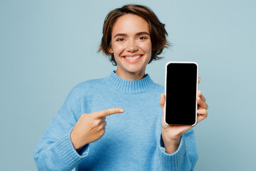 Wall Mural - Young smiling woman wears knitted sweater hold in hand use point index finger on mobile cell phone with blank screen workspace area isolated on plain pastel light blue cyan background studio portrait.