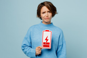 Young weary sad caucasian woman wear knitted sweater look camera hold in hand low battery red card sign isolated on plain pastel light blue cyan background studio portrait. People lifestyle concept.