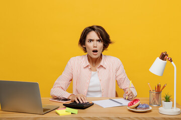 Wall Mural - Young mad employee business woman wear casual shirt sit work at office desk with pc laptop use calculator write down in notebook isolated on plain yellow color background. Achievement career concept.