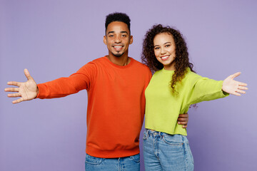 Young couple two friends family man woman of African American ethnicity wear casual clothes together look camera with outstretched hands say you are welcome isolated on pastel plain purple background.