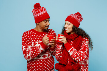 Wall Mural - Merry young couple two man woman in red Christmas sweater Santa hat posing hold in hand cup of tea sip look to each other isolated on plain pastel blue background Happy New Year 2023 holiday concept