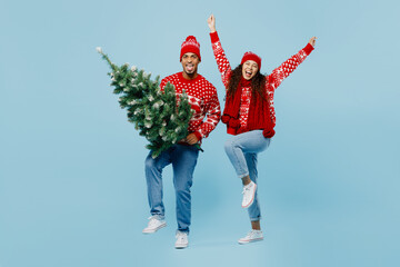 Full body merry young couple two man woman wearing red sweater Santa hat posing hold Christmas tree raise up hands isolated on plain pastel light blue background. Happy New Year 2023 holiday concept.