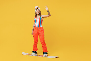 Snowboarder woman wears orange suit goggles mask hat ski costume swimsuit spend extreme weekend waving hand greeting isolated on plain yellow background studio. Winter sport hobby trip relax concept.