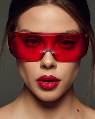 Wall Mural - Close-up portrait of beautiful young woman with red lipstick posing in red plastic glasses over dark grey background. Cosmetology treatment