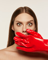 Wall Mural - Portrait of beautiful young woman with straight brown hair and dark makeup gloves over white background. Hand in red rubber glove covering mouth