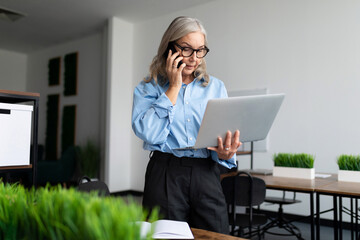 caucasian senior businesswoman speaks on a mobile phone with a laptop in her hands in the office