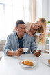 © LIGHTFIELD STUDIOS - blonde woman with cup of coffee smiling near african american boyfriend using smartphone during breakfast