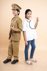 Wall Mural - Happy young indian girl child wearing doctor uniform holding stethoscope and boy kid wearing police costume dress isolated on beige studio background. education concept