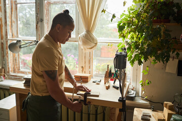 Wall Mural - Side view portrait of tattooed female artist cutting wood while standing by window in cozy workshop, copy space