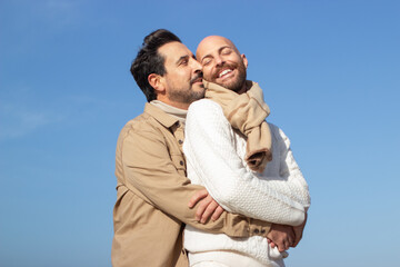 Wall Mural - Happy carefree gays embracing outdoors. Affectionate brunette man whispering in ear of bald boyfriend in scarf against blue sky. Love concept