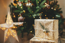 Merry Christmas! Stylish Christmas Gifts At Christmas Tree With Golden Lights. Wrapped Presents With Golden Paper And Fir Branch Under Decorated Tree In Room. Atmospheric Banner, Copy Space